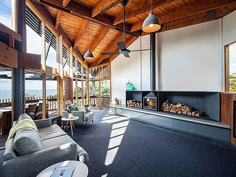 В социальной зоне есть камин с дровяным хранилищем вдоль одной из стен и серым диваном, обращенным к нему. Обеденное пространство расположено за диваном, и две функции тесно связаны, но они выделяются как отдельные области.