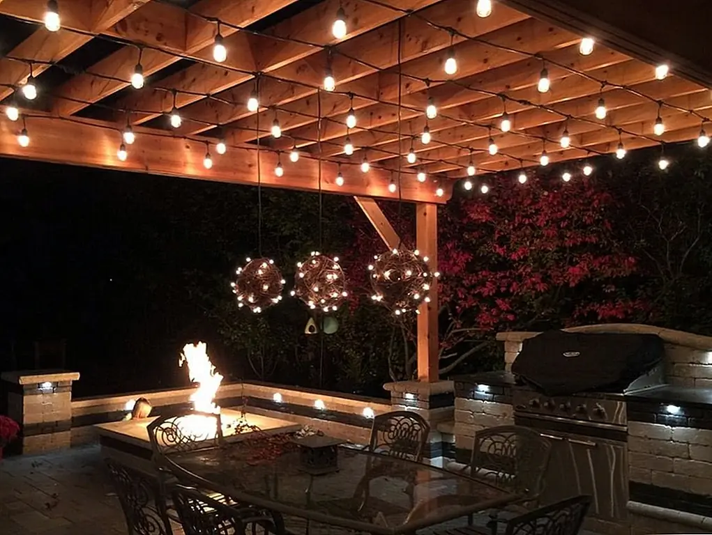 Струнные огни также добавляют окружающий свет, когда они натянуты вокруг висящих деревенских кусков. Изображение: PaveStone Brick Paving