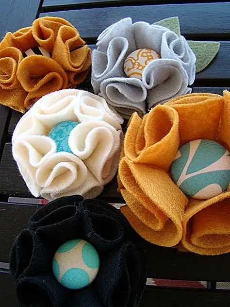 Этот проект немного проще, но так же хорош. Принадлежности, необходимые для одного цветка, - это полный кусочек готового войлока, кнопка, горячий пистолет для клея и булавка. Используйте чашку или что-то похожее на круги кругов на войлоке. Вам нужно шесть кругов на цветок для лепестков и меньший для базы. Сложите каждый круг пополам, а затем сместите угол, чтобы сделать треугольник. Сложите другой угол назад на S, а затем положите немного клея с каждой стороны, чтобы удерживать его на месте. Центрируйте треугольники на базовом круге и прикрепите их клеем. Затем добавьте кнопку в центр. {Найдено на befickle}.